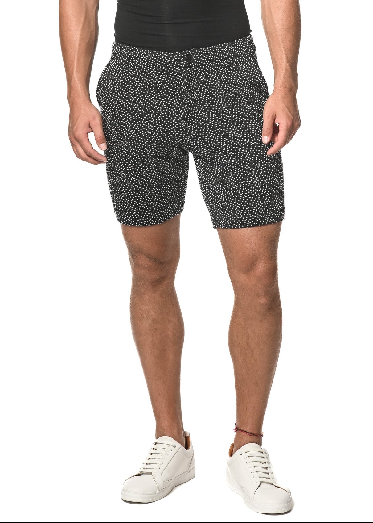 A man wearing black and white dot patterned shorts with a stretch waistband, belt loops, side pockets, and button-through welt pockets, paired with a black V-neck t-shirt and white sneakers.